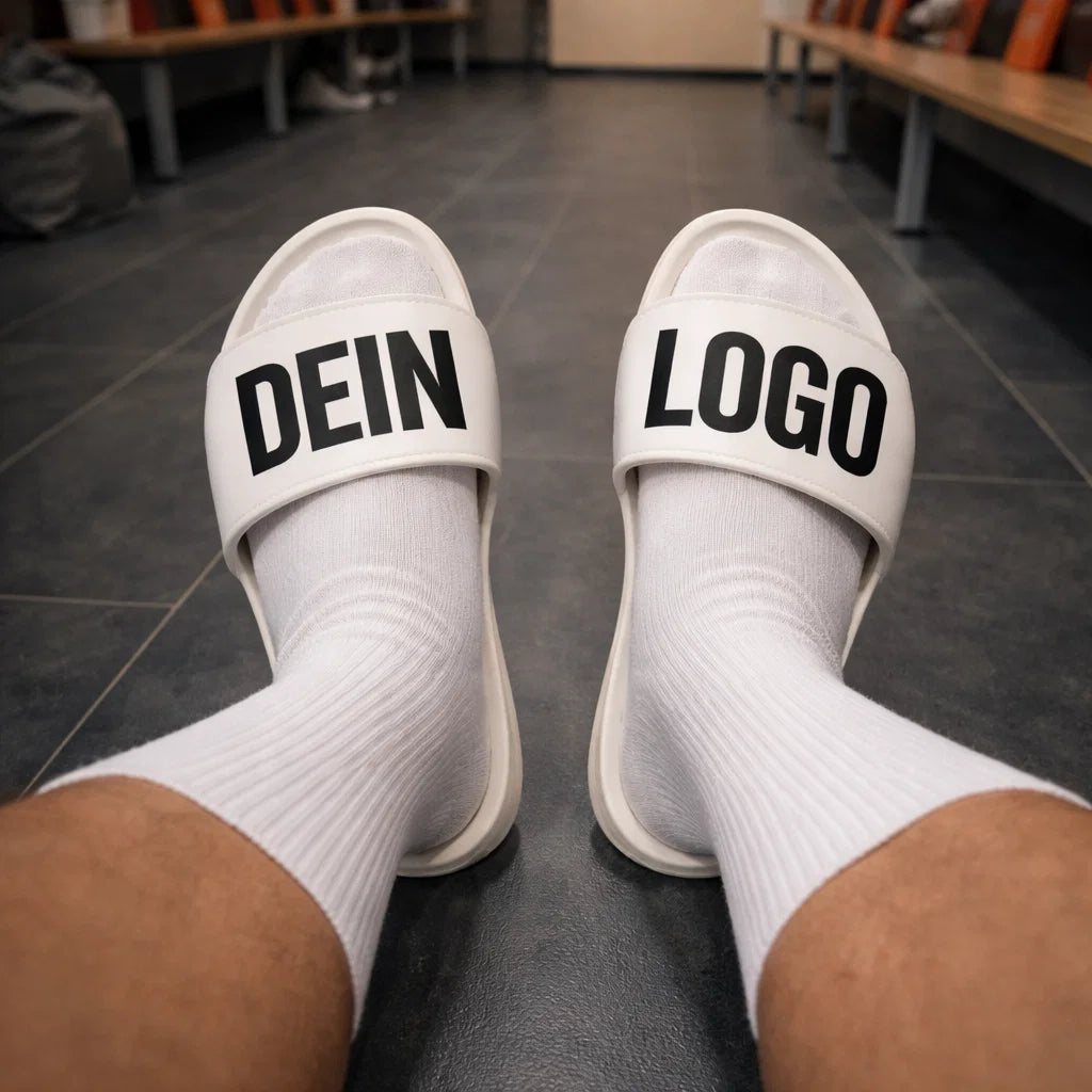 Adiletten in der Kabine getragen, weißer Riemen mit „DEIN LOGO“ vor Spinden im Hintergrund, Adiletten personalisieren