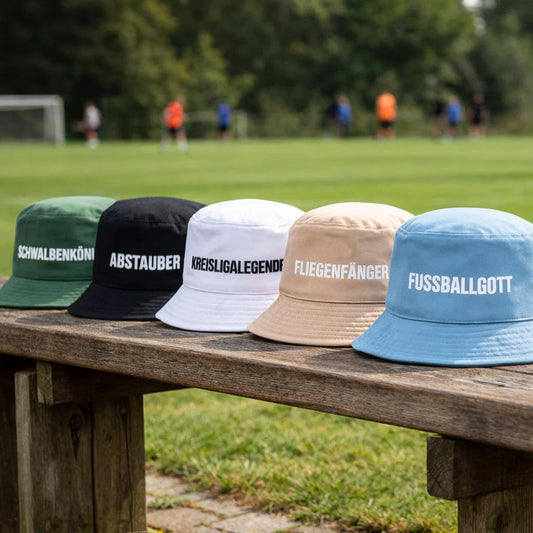 Mehrere Fischerhüte mit Spruch in Beige, Blau, Grün, Schwarz und Weiß auf einer Bank am Fußballplatz