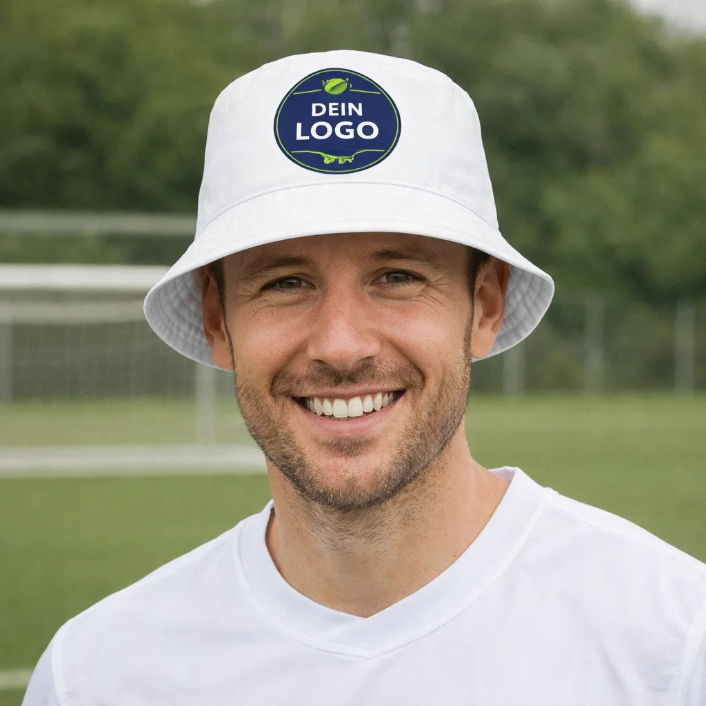 Weißer Fischerhut mit „DEIN LOGO“-Patch, getragen von Mann auf Sportplatz – Fischerhut bedrucken